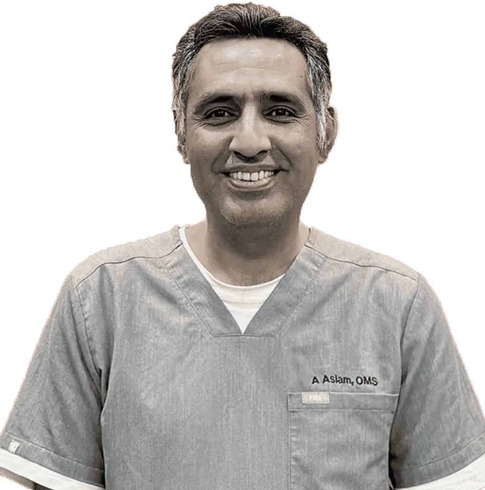 A middle-aged man, likely a doctor and medical professional, with short graying hair, smiling and wearing light-colored medical scrubs. A small name badge is visible on his chest.