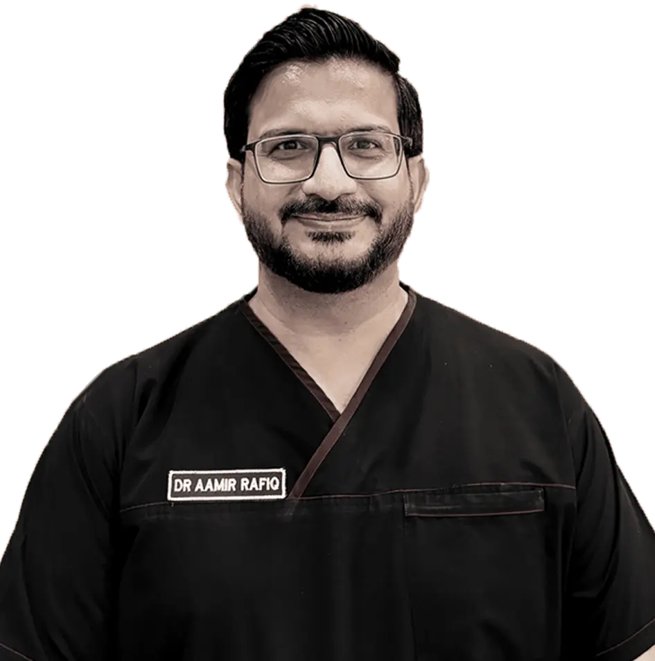 A headshot of a smiling man, likely a doctor or surgeon, with a beard and glasses, wearing dark medical scrubs that have a name tag reading "DR AAMIR RAFIQ" visible on the chest.