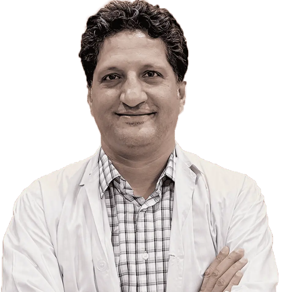 A headshot of a smiling man, likely a doctor or medical professional, with curly dark hair. He is wearing a white lab coat over a light-colored plaid shirt and has his arms crossed.