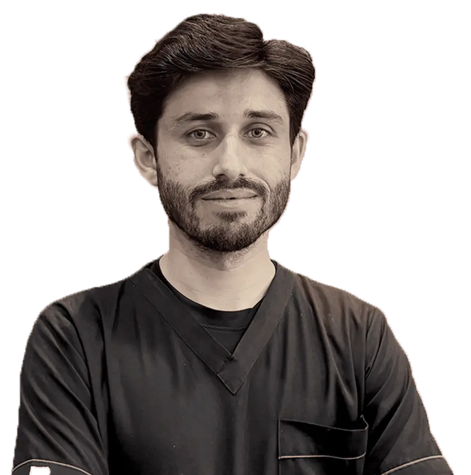 A headshot of a young man, likely a medical professional, with dark hair and a beard, looking directly at the camera. He is wearing dark medical scrubs. The image is in black and white or a sepia tone.